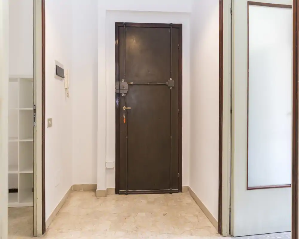 Simple entry hallway with a sturdy dark metal door, intercom on the wall and light marble floor — a clear view of the apartment entrance.