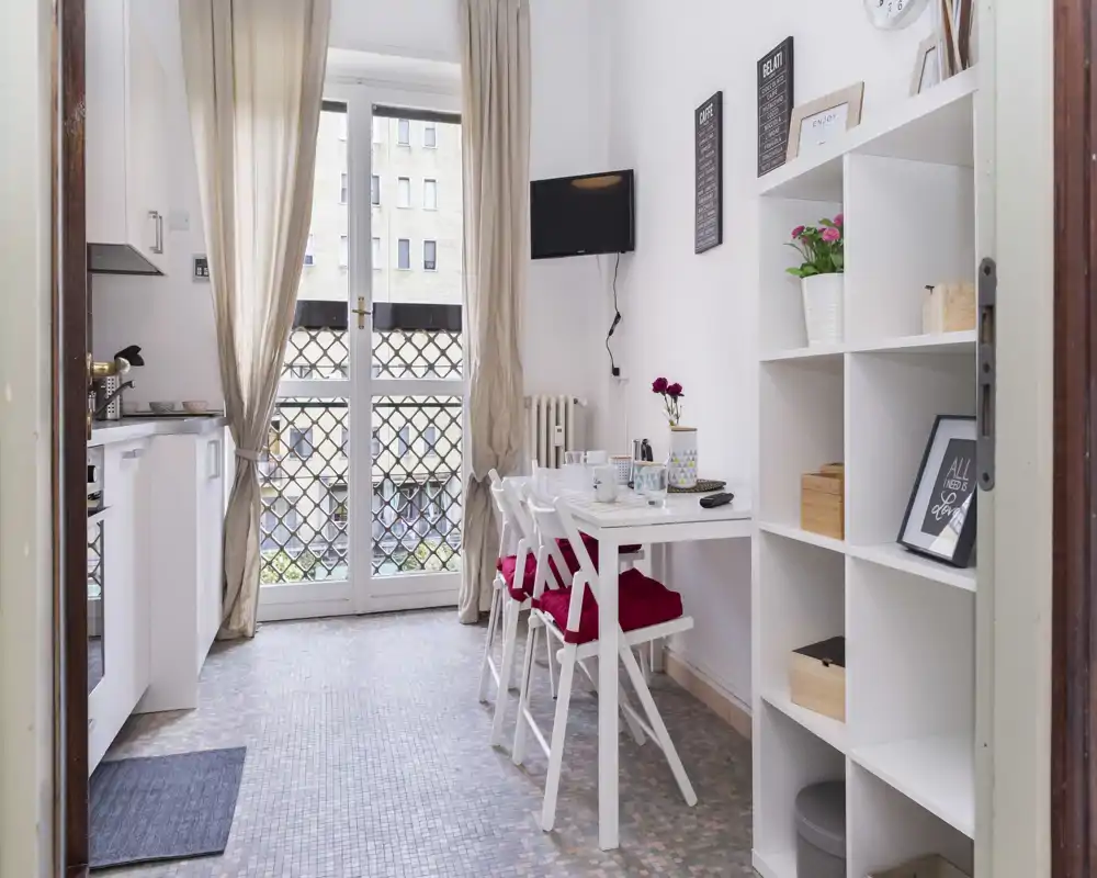 Compact, bright kitchenette with a small dining table for four, white cabinetry and a large glass door providing natural light and a pleasant city view.