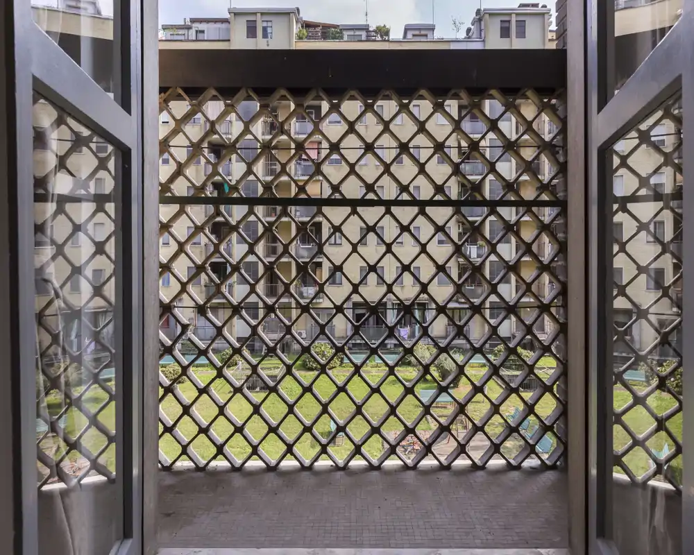 Small balcony view looking out through a decorative metal grille onto a communal courtyard with grass and neighboring apartments.