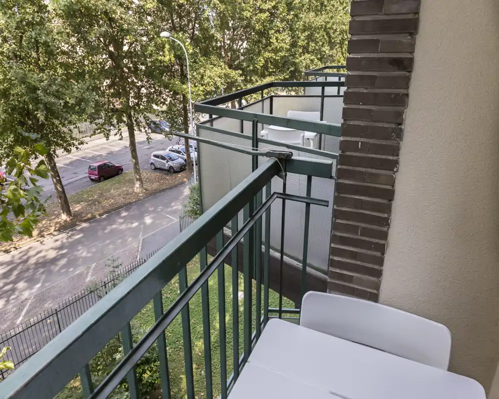 Sunny private balcony with a small table and chairs, overlooking a tree-lined street — ideal for morning coffee or relaxing outdoors.