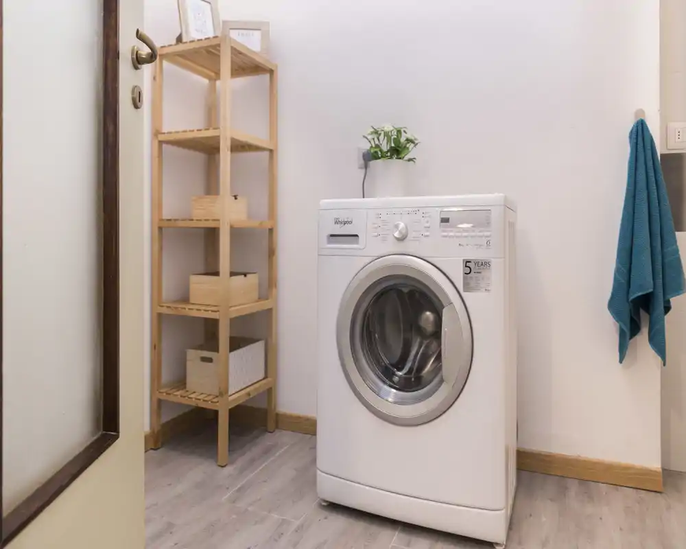 A compact laundry/utility area featuring a freestanding front-load washing machine, wooden shelving for storage and a hanging towel — bright and neat.
