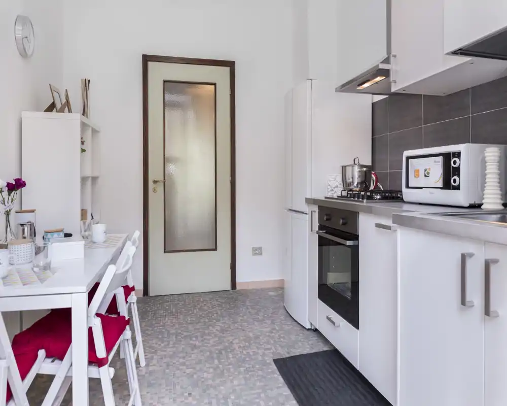 Bright, compact kitchen with white cabinets, oven, microwave and a small dining table with red cushions; neat, functional layout ideal for a city apartment.