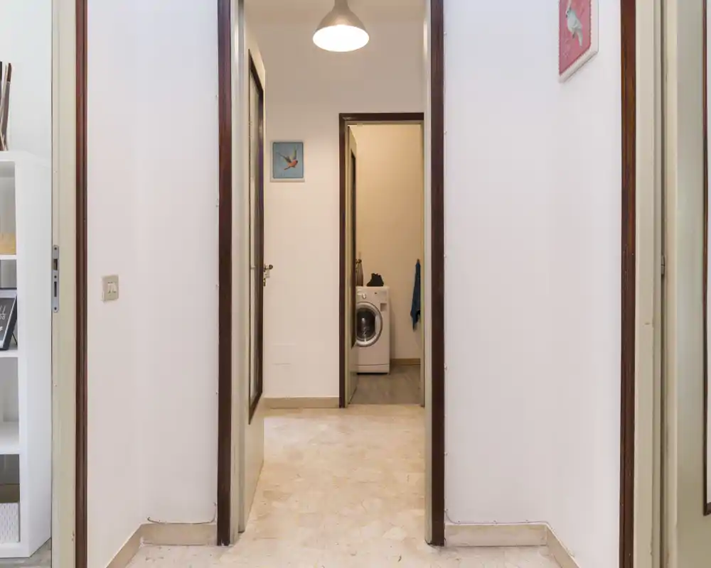 Bright, simple hallway/entrance with tiled floor leading to an inner room; clean walls and pendant light provide a neutral, welcoming arrival space.