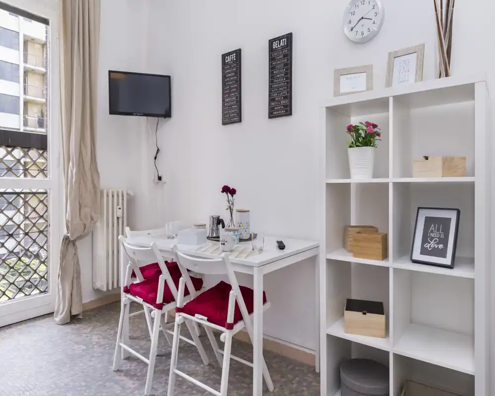 Small, bright dining nook with a white table, three chairs with red cushions, decorative shelving and natural light from a window — a cozy spot for meals.