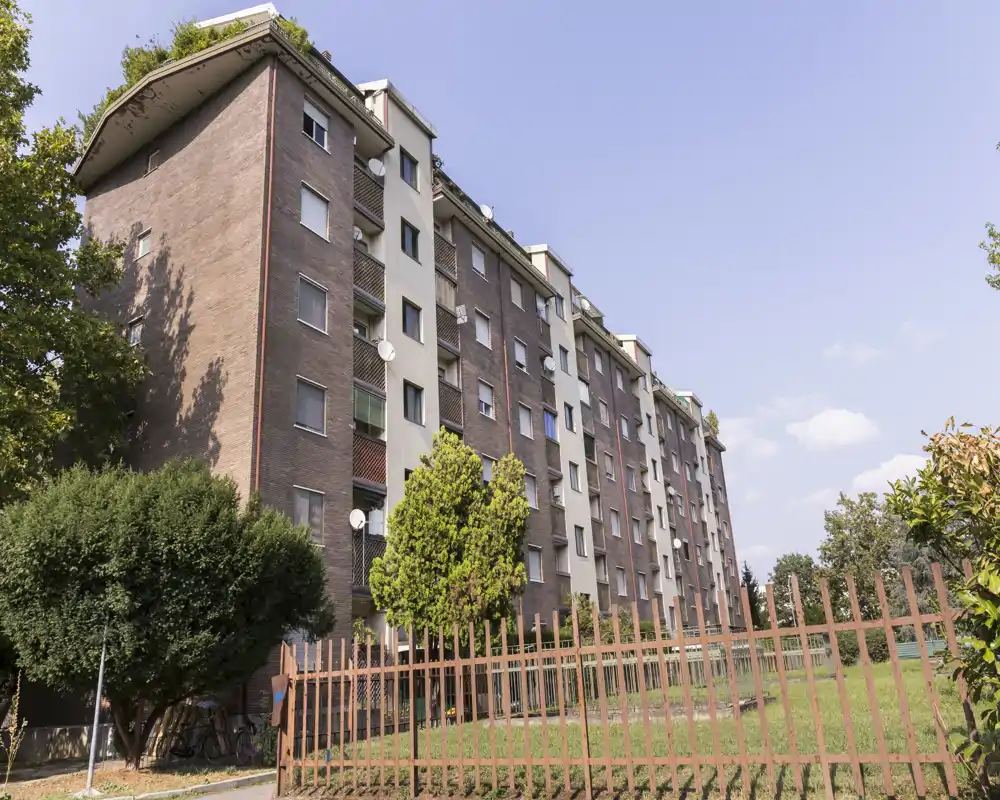 Exterior view of a multi-story residential building with a fenced grassy courtyard and mature trees, photographed in clear daylight.