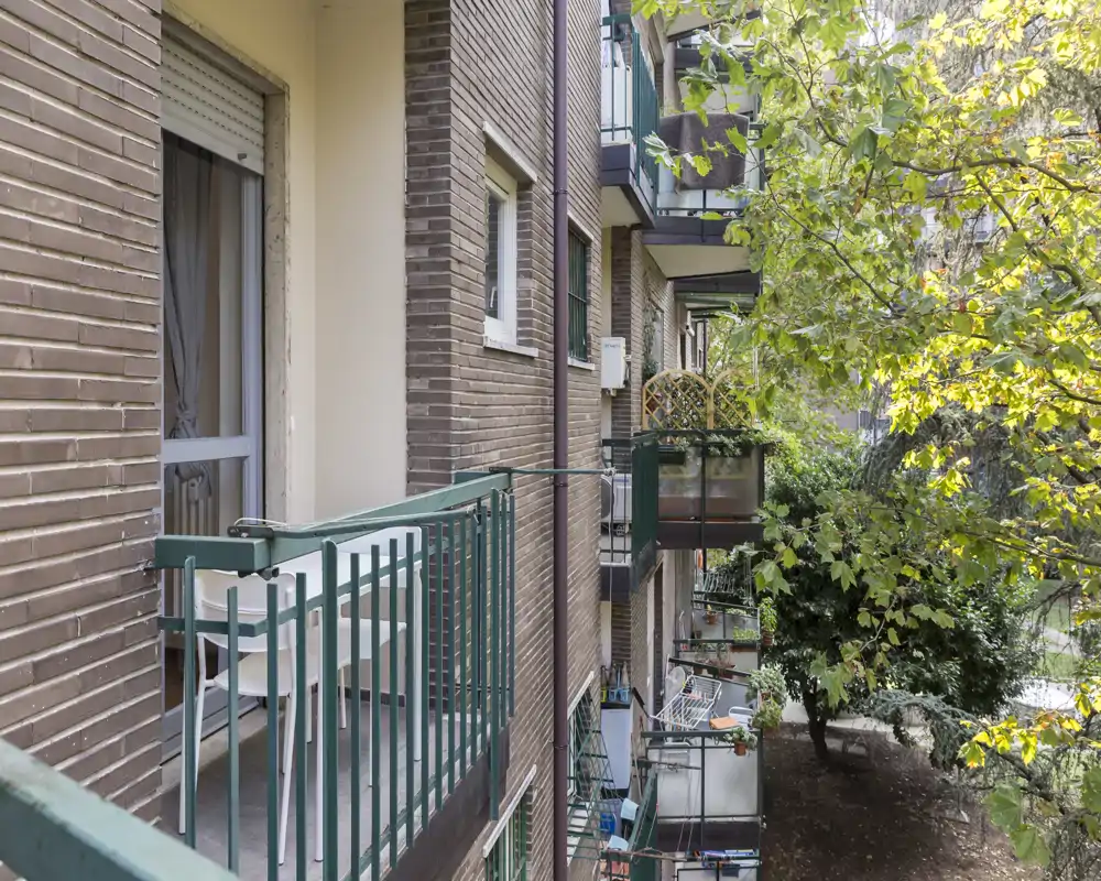 Small apartment balconies overlooking a leafy courtyard; green railings and potted plants with pleasant natural light.