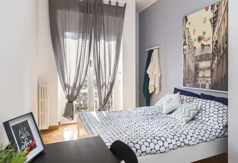 Chambre lumineuse et soignée avec un lit double, une couette à motifs, des coussins décoratifs et une grande fenêtre aux rideaux légers laissant passer une douce lumière naturelle.