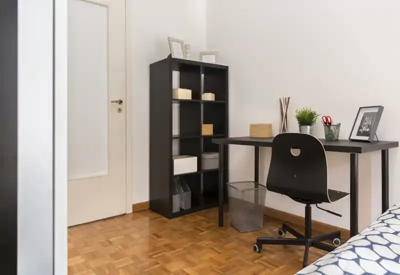 Coin de chambre lumineux et soigné avec un bureau simple, une chaise pivotante et une étagère; une partie du lit est visible sur le bord droit de l'image.