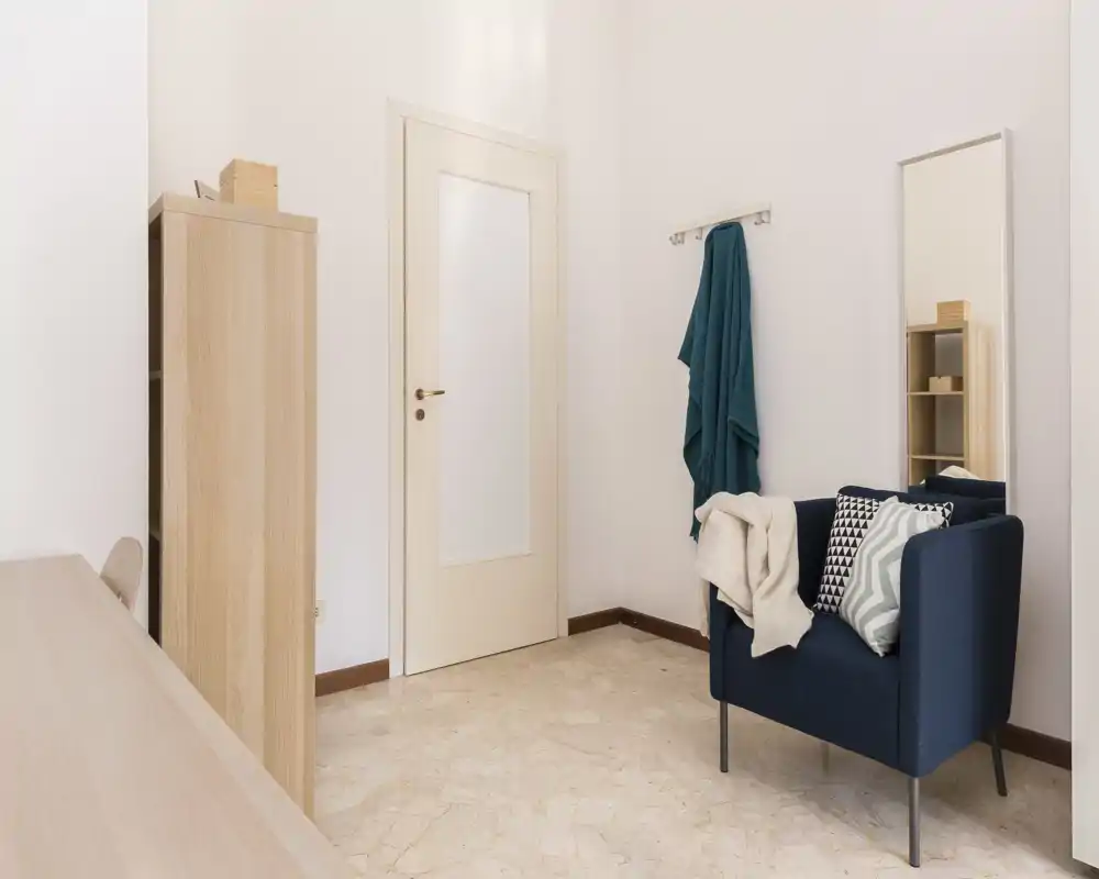 Small, well-kept living area with a comfortable armchair, cushions and a coat hook; clean neutral tones create a calm, functional corner ideal for reading or resting.