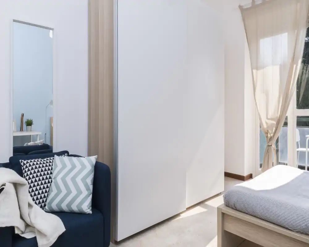 Bright, modern bedroom with a wooden bed frame, striped bedding and soft natural light from a balcony window; decorative cushions and a cozy throw create a welcoming atmosphere.