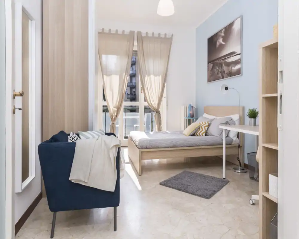 Bright, well-staged bedroom with a wooden bed, soft textiles and natural light from a large window; a cozy armchair and desk complete the space.