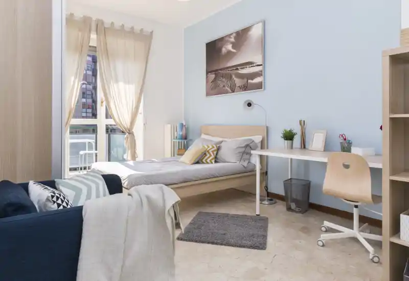 Camera luminosa e ben arredata con letto singolo, scrivania e accesso al balcone. Pareti azzurre, cuscini comfortevoli e luce naturale rendono l'ambiente accogliente, ideale per uno studente o una persona sola.