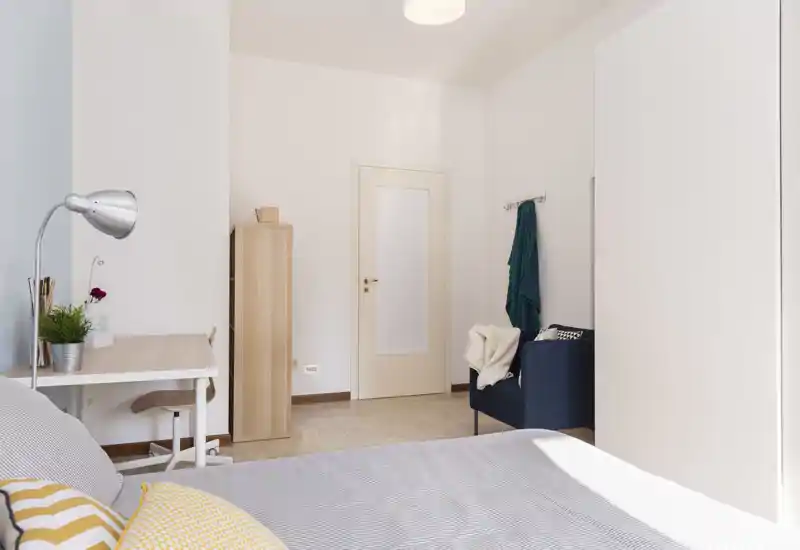 Camera da letto luminosa e minimal con letto ben rifatto, una scrivania compatta e una poltrona accogliente vicino alla porta — ideale per una persona o soggiorni brevi.