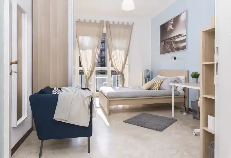 Camera da letto luminosa e ben arredata con letto in legno, tessili morbidi e luce naturale da una grande finestra; una poltrona accogliente e una scrivania completano l’ambiente.