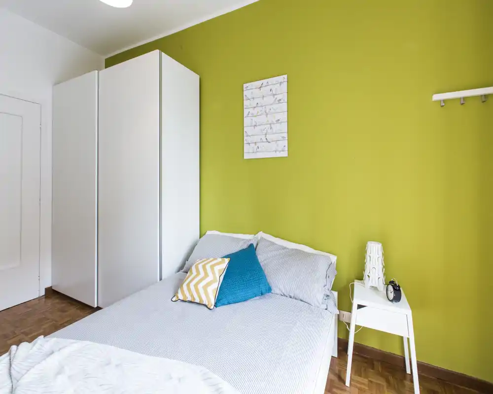 Chambre lumineuse et moderne avec lit double devant un mur d'accent vert vif, coussins décoratifs, armoire blanche et table de chevet minimaliste avec lampe — idéale pour un séjour confortable.