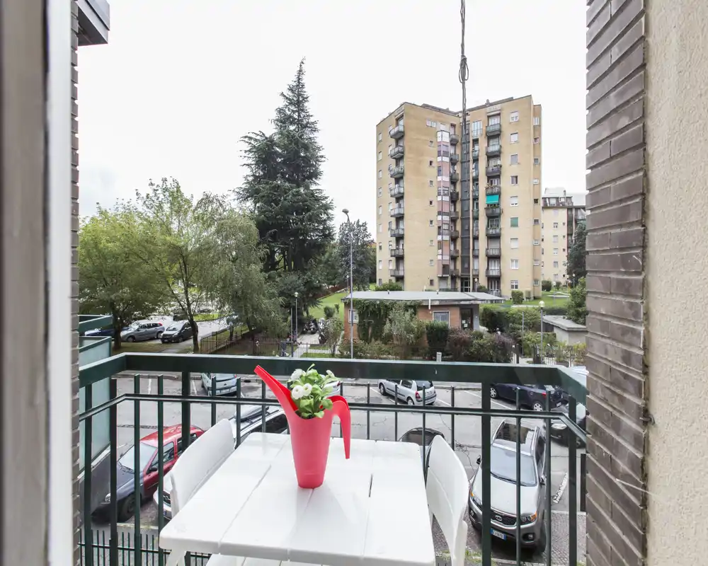 Balcon ensoleillé avec une petite table, deux chaises et un arrosoir rouge utilisé comme pot; vue sur une cour résidentielle et des immeubles — idéal pour le café du matin.