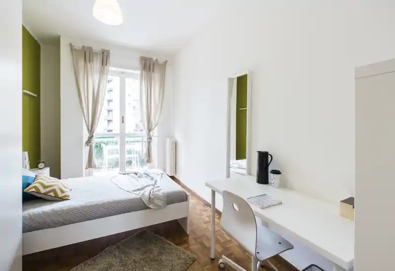 Chambre moderne et lumineuse avec un lit confortable, un bureau et une grande fenêtre donnant sur un balcon. Tons neutres et mur vert en accent, avec des textiles doux pour une ambiance apaisante et accueillante.