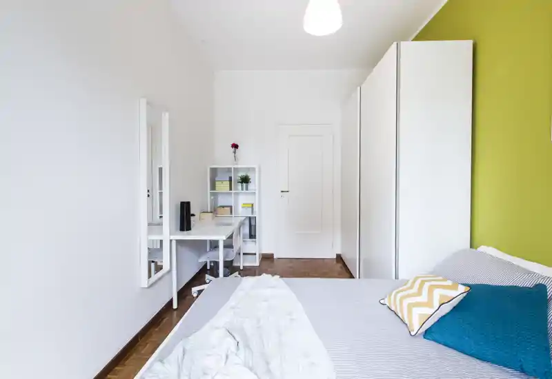 Chambre lumineuse et moderne avec un lit double confortable au premier plan, coussins décoratifs et un mur vert contrastant ; bureau et rangement à l'arrière pour un coin travail soigné.