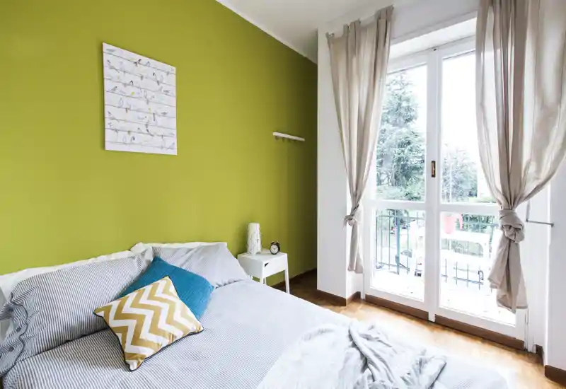 Chambre lumineuse et cosy avec un lit double adossé à un mur vert, coussins décoratifs, une petite table de chevet blanche et une grande fenêtre/porte laissant entrer une belle lumière naturelle.