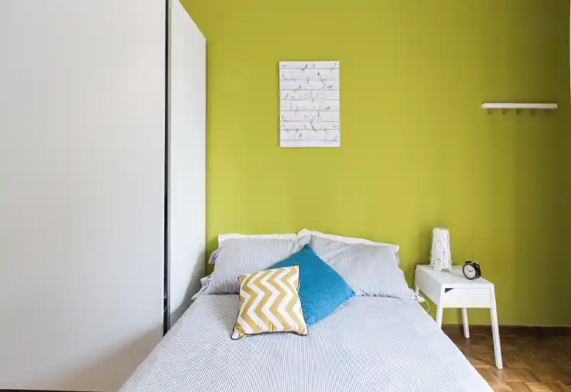 Chambre lumineuse et bien mise en scène avec un lit double, coussins colorés et une table de chevet blanche ; mur vert vif et armoire à miroir apportent style et lumière.