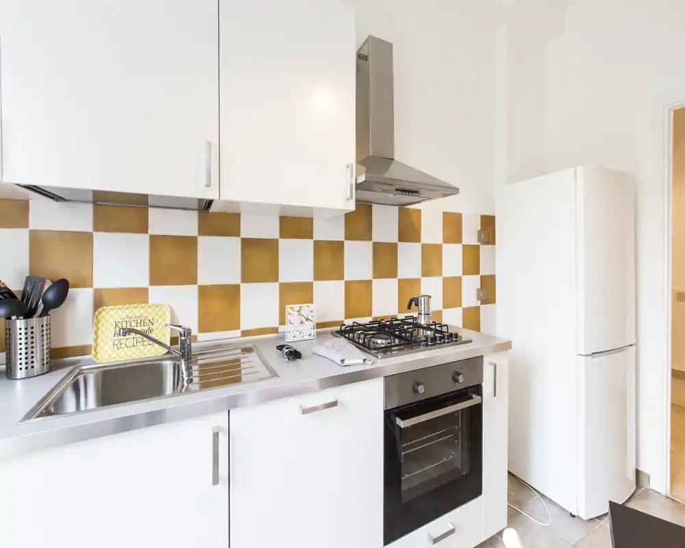 Cuisine moderne et lumineuse avec placards blancs, évier inox, plaque à gaz et four, et un carrelage à damier qui apporte de la chaleur.