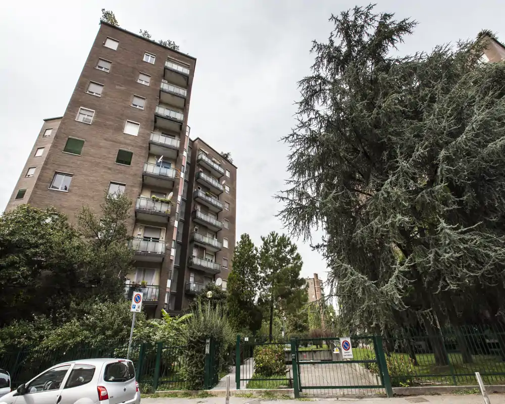 Vue extérieure d'un immeuble résidentiel avec balcons et une cour verdoyante clôturée ; arbres matures et stationnement de rue visibles sous un ciel couvert.
