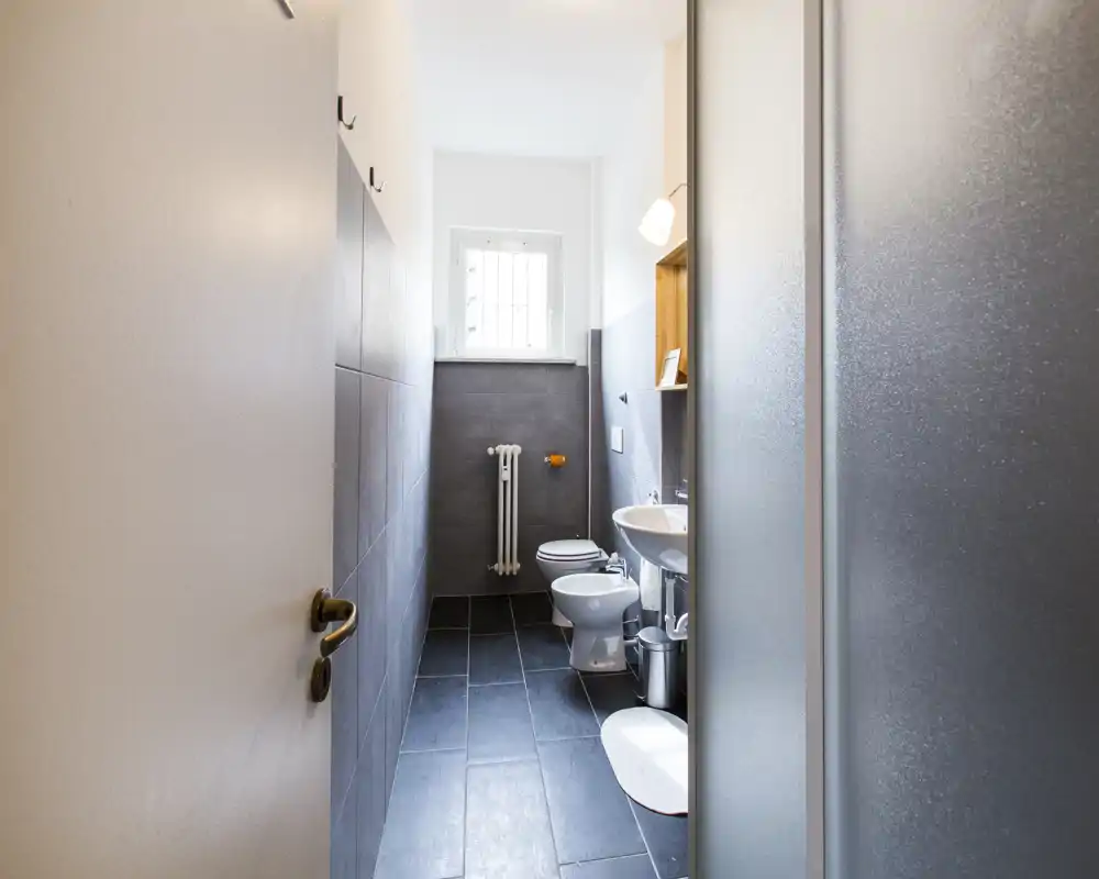 Salle de bains moderne et compacte avec carrelage sol foncé, sanitaires blancs (toilettes, bidet et lavabo), une petite fenêtre et une paroi de douche en verre dépoli. Espace propre et bien éclairé, idéal pour les locations courte durée.