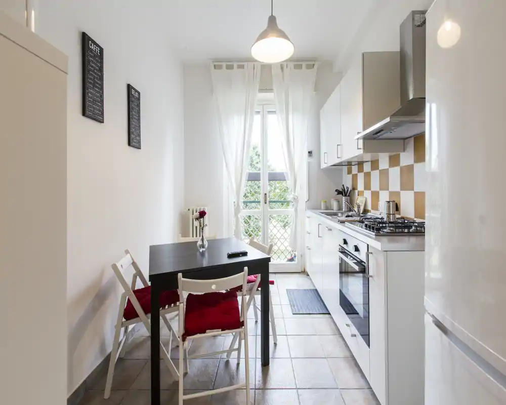 Cuisine moderne et lumineuse en enfilade avec placards blancs, four encastré et plaque à gaz, ainsi qu'une petite table avec chaises à coussins rouges près d'une porte ensoleillée.