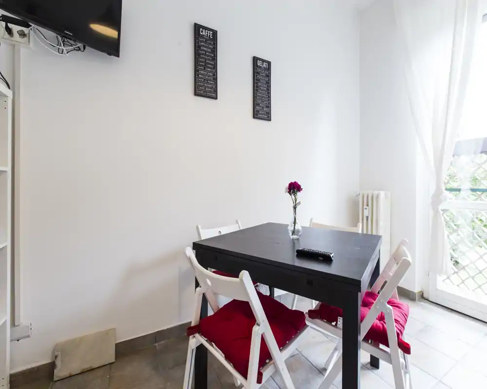 Coin repas compact avec une table carrée noire et quatre chaises pliantes blanches équipées de coussins rouges, lumière naturelle vive et un vase avec une fleur pour une touche cosy.