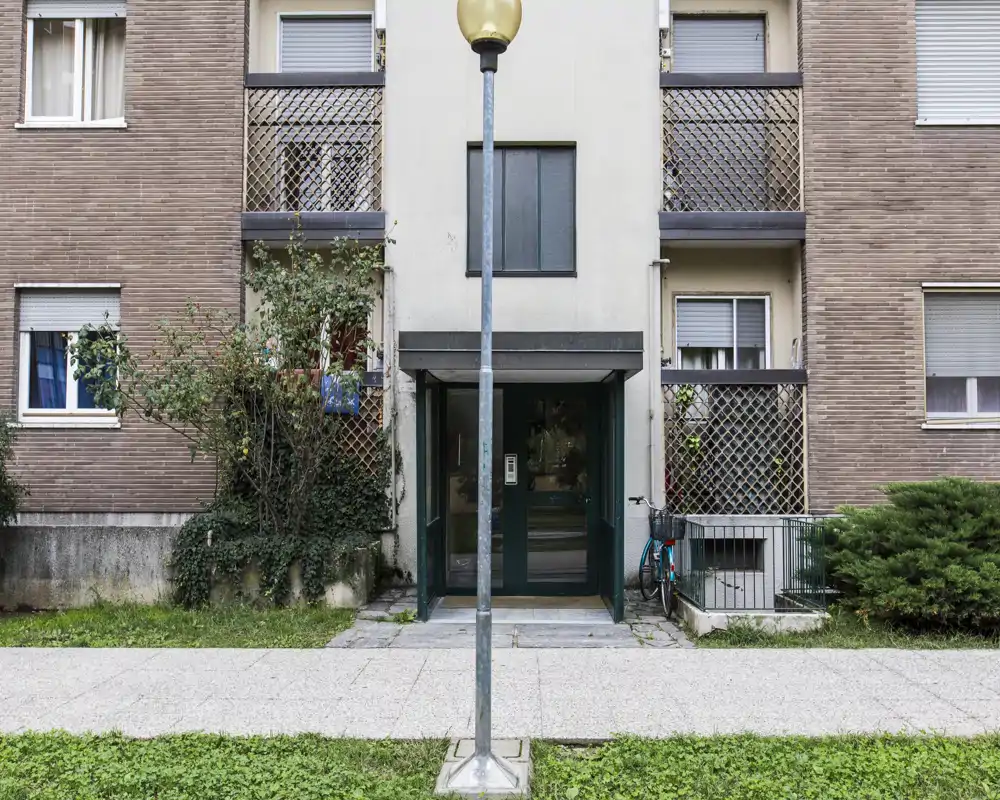Entrée au rez-de-chaussée d'un immeuble résidentiel avec portes vitrées doubles, interphone et balcons latéraux; un lampadaire et un vélo sont visibles au premier plan.