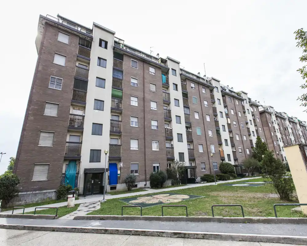 Vue extérieure d'un immeuble résidentiel avec pelouse commune et allée pavée à l'avant, montrant plusieurs entrées et balcons.
