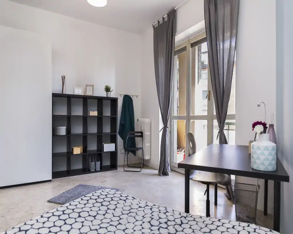 Chambre lumineuse et minimaliste avec couvre-lit à motifs, un bureau près de la fenêtre et une étagère; de grandes portes de balcon apportent une belle lumière naturelle.