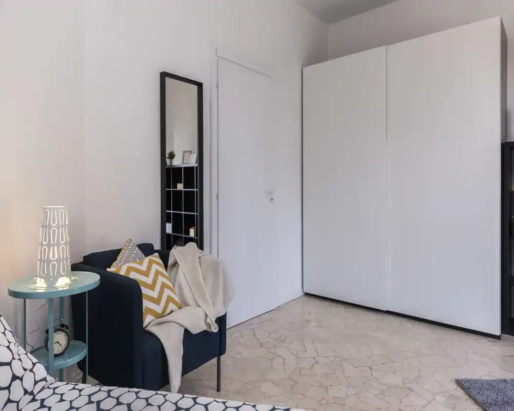 Coin chambre chaleureux et moderne avec couvre-lit à motif, fauteuil décoré de coussins et grande armoire. Murs neutres et sol carrelé apportent une atmosphère lumineuse et apaisante.