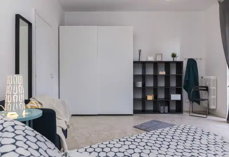 Chambre lumineuse et minimaliste avec lit à motifs au premier plan, grand placard blanc et étagères ouvertes contre le mur. Textiles doux, lampe de chevet et tons neutres pour une atmosphère moderne et apaisante.