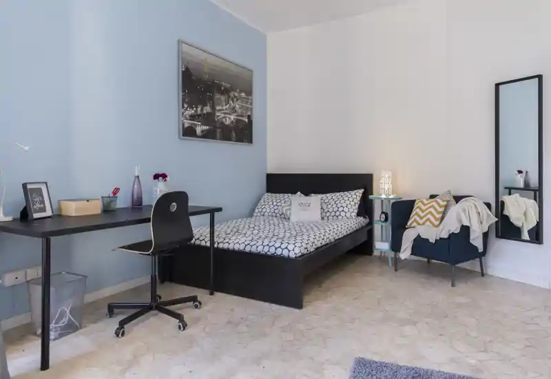 Chambre moderne et lumineuse avec lit double, literie à motifs, bureau avec chaise et fauteuil confortable avec lampe — idéale pour se reposer et travailler.