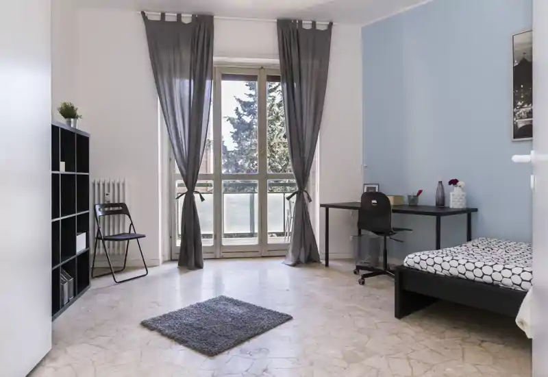 Chambre lumineuse et minimaliste avec un lit simple, un bureau et une grande fenêtre/porte-fenêtre donnant sur le balcon qui apporte beaucoup de lumière naturelle; étagères simples et petit tapis.