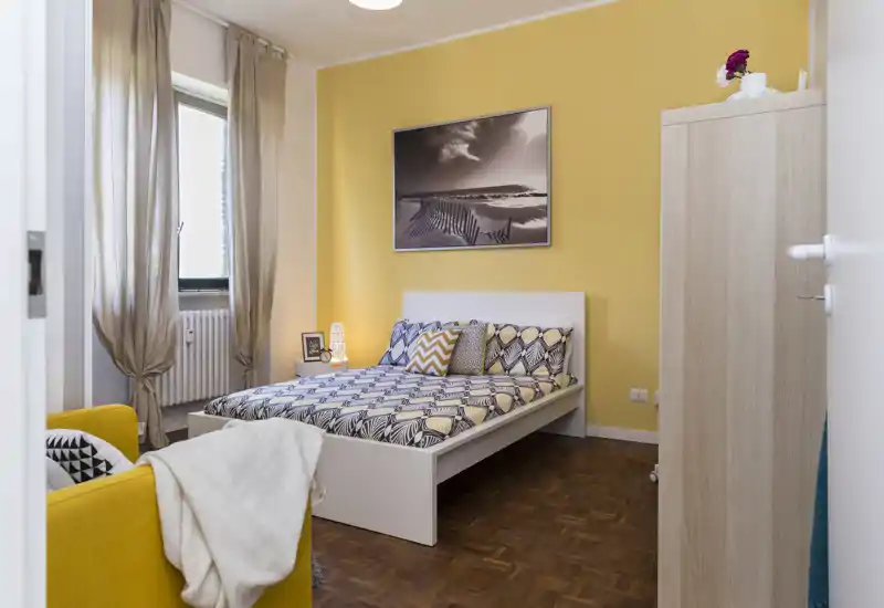 Chambre lumineuse et bien décorée avec un lit double, une literie à motifs et un mur d'accent jaune. La lumière naturelle et la décoration soignée créent une atmosphère chaleureuse et accueillante.