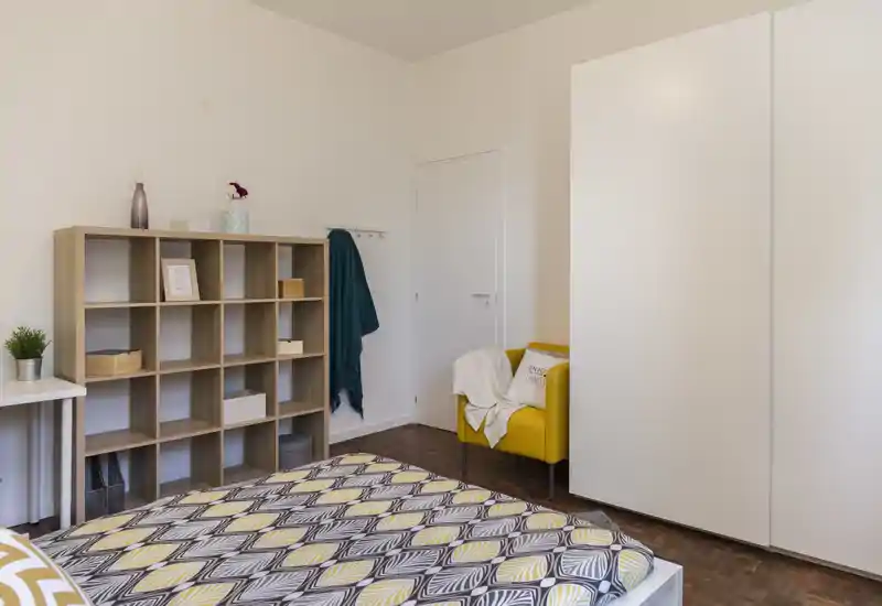 Chambre lumineuse et moderne avec un lit double à motif au premier plan, étagères ouvertes, une armoire blanche et un fauteuil jaune avec un plaid chaleureux.