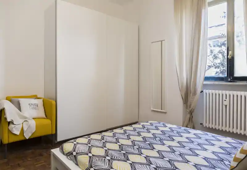 Chambre moderne et lumineuse avec une couette à motif, une grande armoire blanche et un fauteuil moutarde près de la fenêtre — chaleureuse, bien éclairée et parfaitement mise en scène pour une annonce.