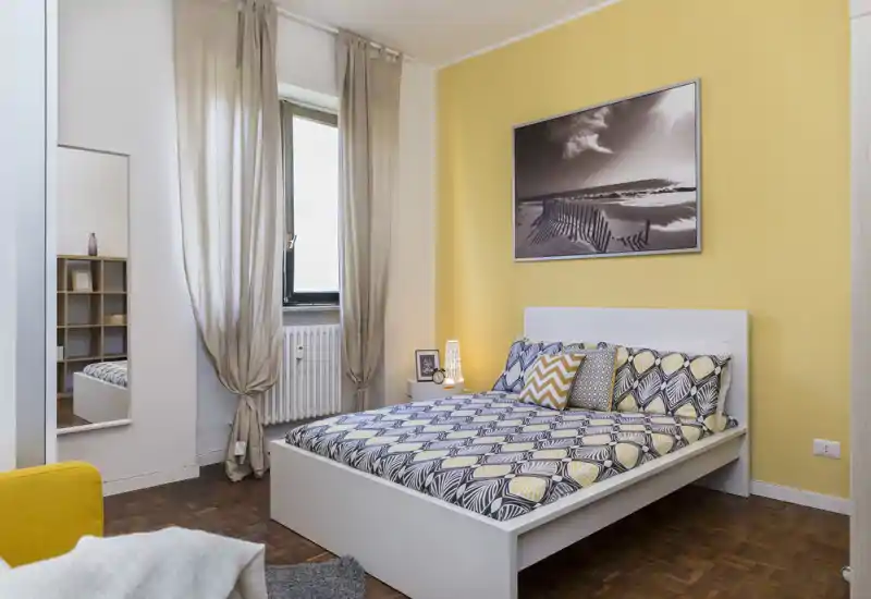 Chambre lumineuse et soignée avec un lit double à motifs, coussins assortis et un mur jaune chaleureux; la lumière naturelle et la décoration créent une atmosphère accueillante.