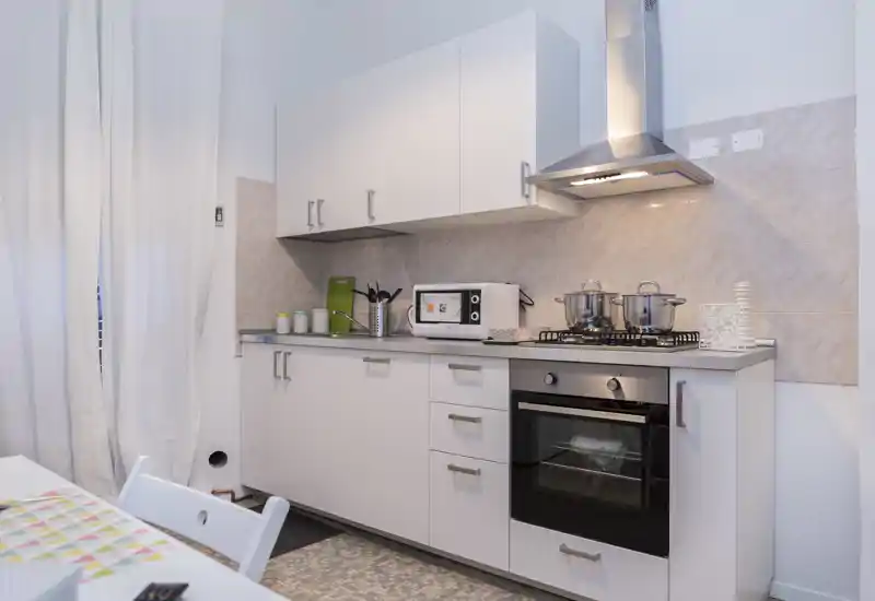 Cuisine compacte et moderne avec meubles blancs, four à gaz, hotte et micro-ondes sur le plan de travail ; lumineuse, propre et prête à cuisiner.