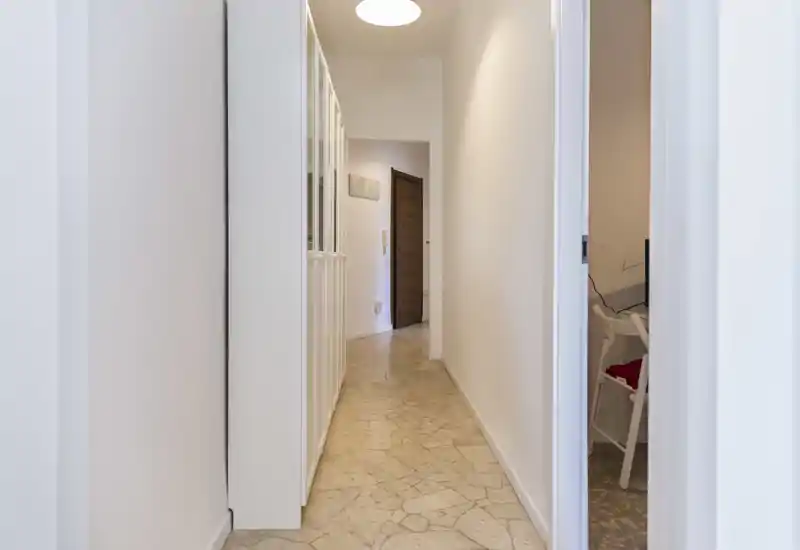 Couloir d'entrée lumineux et étroit avec sol imitation marbre, murs blancs et rangements encastrés sur un côté ; espace propre et bien éclairé menant à la porte intérieure.