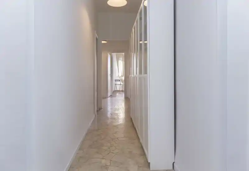 Couloir étroit et lumineux aux murs clairs et au sol carrelé, menant à d'autres pièces ; entrée minimale avec rangement intégré sur la droite.