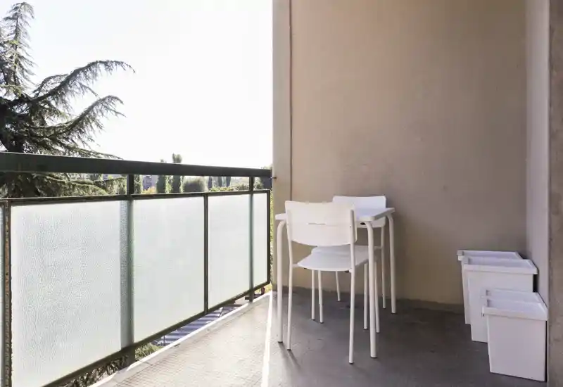 Balcon ensoleillé avec une petite table blanche et chaises empilées, garde-corps en verre dépoli et vue sur les arbres — un coin extérieur compact pour le café du matin.