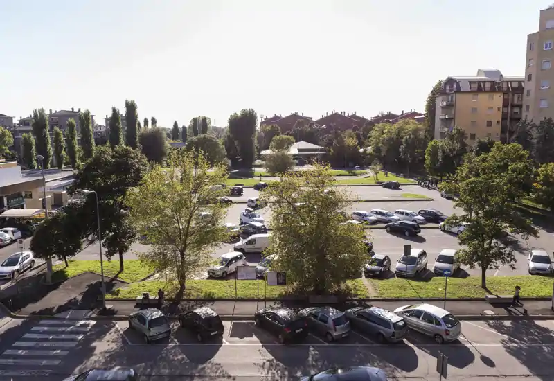 Vue extérieure urbaine ensoleillée montrant un parking, des arbres environnants et des immeubles à proximité — idéale pour mettre en valeur le stationnement extérieur et les espaces verts.