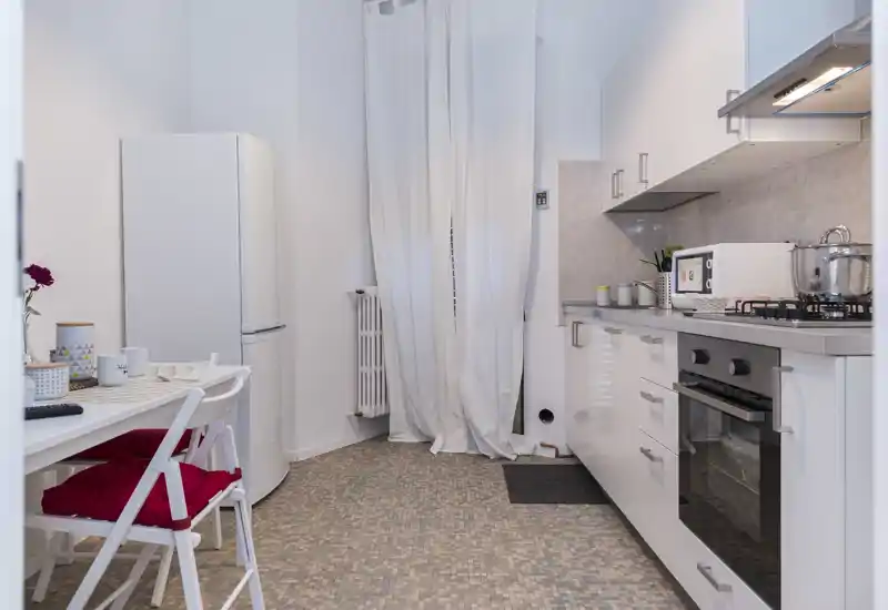 Cuisine moderne et lumineuse avec meubles blancs, four encastré, plaque à gaz et petite table à manger avec chaises. L’espace est propre et décoré avec quelques accessoires, idéal pour un appartement citadin.