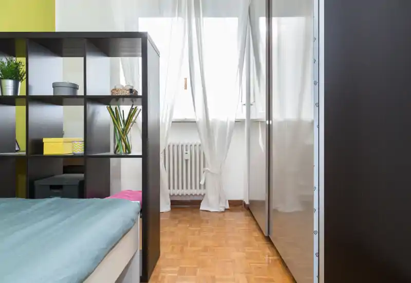 Chambre compacte et lumineuse avec un lit bas partiellement visible derrière une étagère-cloison, parquet et armoire sur le mur de droite. La lumière naturelle traverse des voilages, offrant une atmosphère fraîche et soignée.