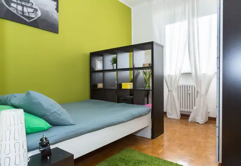 Chambre moderne et lumineuse avec un lit double confortable, coussins décoratifs et une étagère design servant de séparateur; la lumière naturelle met en valeur le mur vert.