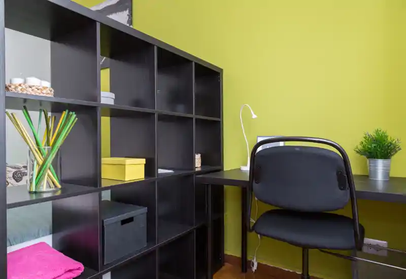 Coin chambre compact avec étagères et petit bureau : une bibliothèque modulaire noire sépare partiellement l'espace lit d'un poste de travail avec chaise, lampe et plante.