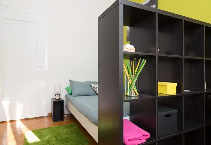 Chambre chaleureuse et lumineuse avec un lit simple partiellement visible derrière une étagère ouverte moderne ; linge frais, coussins décoratifs et tapis vert créent une atmosphère accueillante.